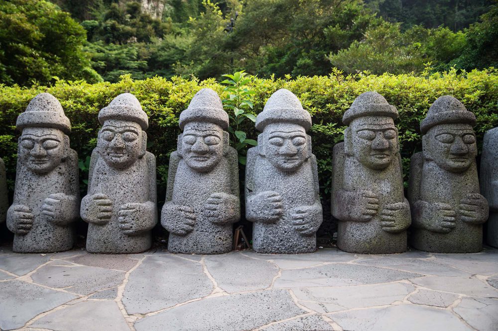 A line of Dol Hareubang statues in a green surrounding on Jeju Island.